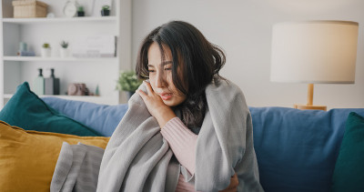 生病的年轻亚洲妇女头痛坐在沙发上的毯子在客厅在家里。