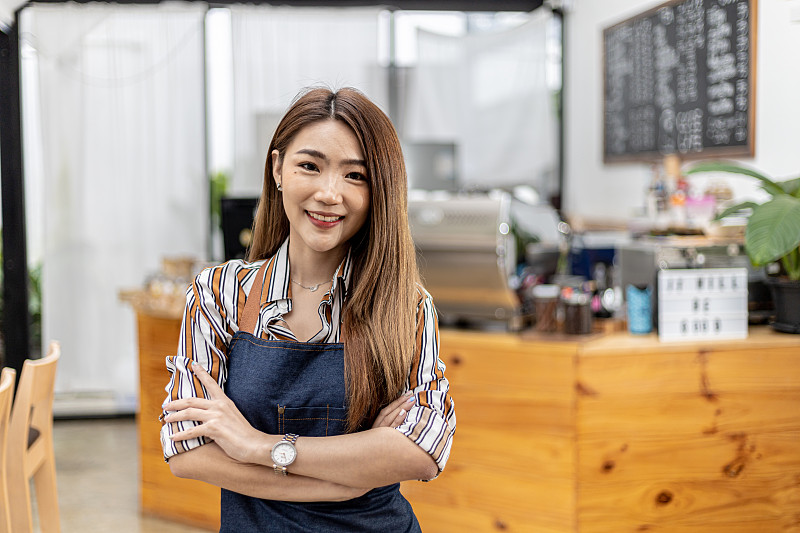 一名美丽的亚洲女子穿着围裙站在一家咖啡店里，她拥有一家咖啡店，经营一家餐饮概念店。由女商人管理的商店