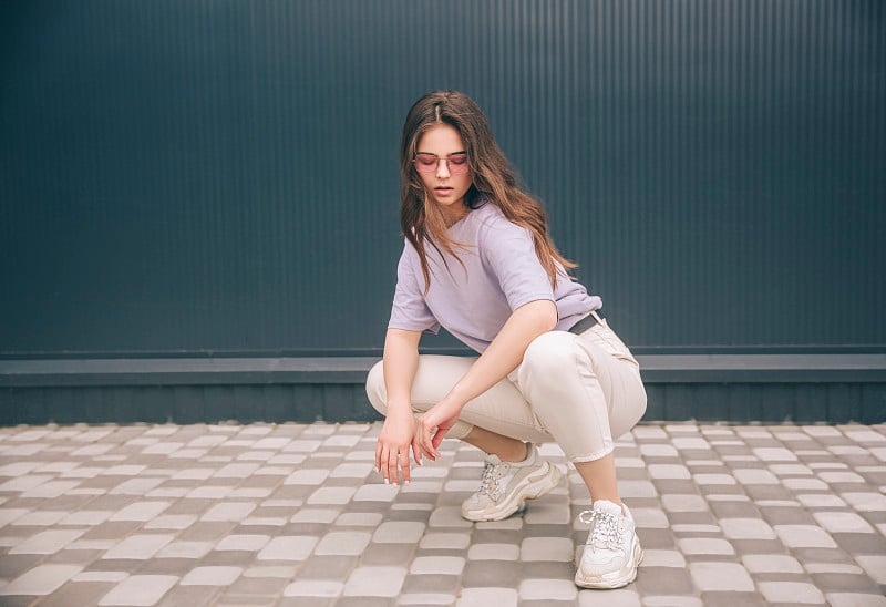 年轻时髦的女人孤立在灰色的蓝色背景。漂亮的女孩穿着紫色的衬衫和白色的裤子，蹲坐着看着镜头。时髦的位置