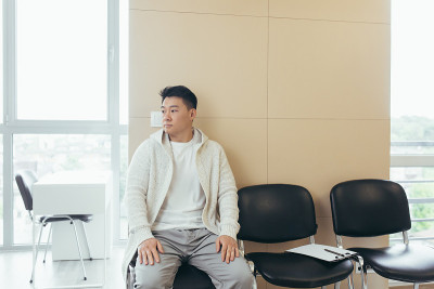 一个年轻的亚洲人坐在候诊室的走廊里等待面试或会议。学生或参赛者在接待处考试或人事。男病人在医院诊所办