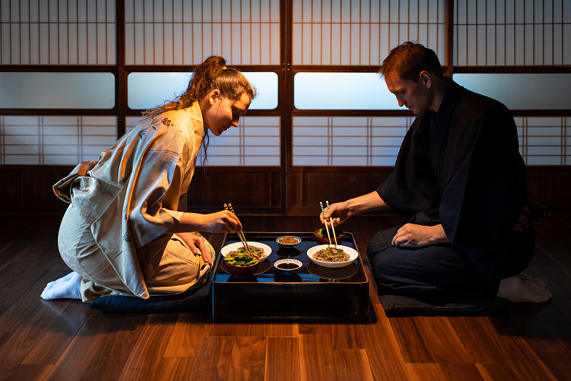 一对年轻夫妇坐在地板上抱着枕头，吃着日式怀石料理菜，把荞麦面、纳豆、毛豆和酱油放在桌子上，由正司滑动