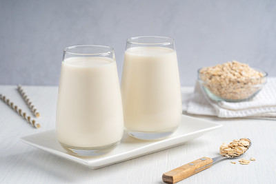 两杯不含素食乳制品的燕麦牛奶放在白色木制背景的盘子里，碗里有燕麦片、勺子和纸吸管