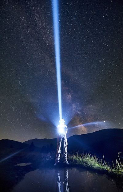 在山区的夜晚，穿着太空服的宇航员借助灯笼将一束光射向天空