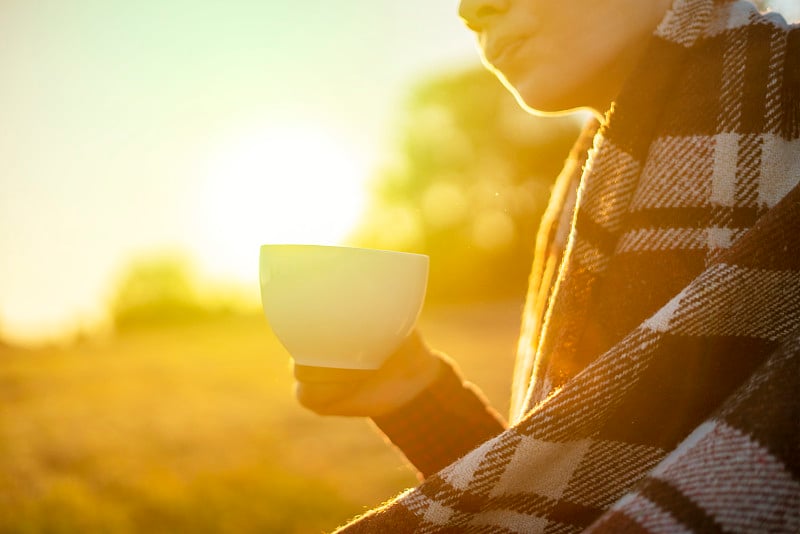 在日落时分，年轻的冻僵的男人在温暖的毯子里坐在田野里喝茶