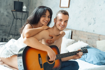 混血夫妇。年轻的男人弹吉他，女人抱着他坐在家里的床上，看着镜头兴高采烈