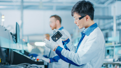 高科技工厂:质量控制工程师拾起并视觉检查智能电子设备可能的损坏。采用表面贴装取放技术的PCB后台装配