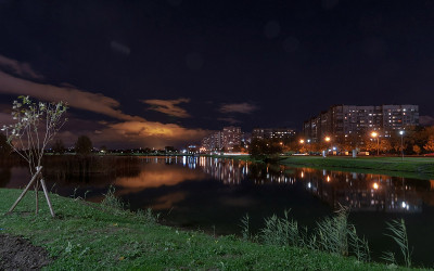 夜晚的城市景观-高层住宅与明亮的窗户在池塘岸边，黑暗的天空与云朵和倒影在水中