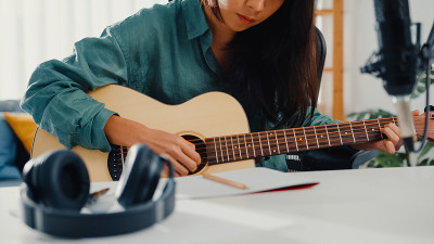快乐的亚洲女性作曲家弹奏原声吉他，从智能手机上听歌曲，思考，写笔记，在纸上写歌词，在家庭工作室的客厅