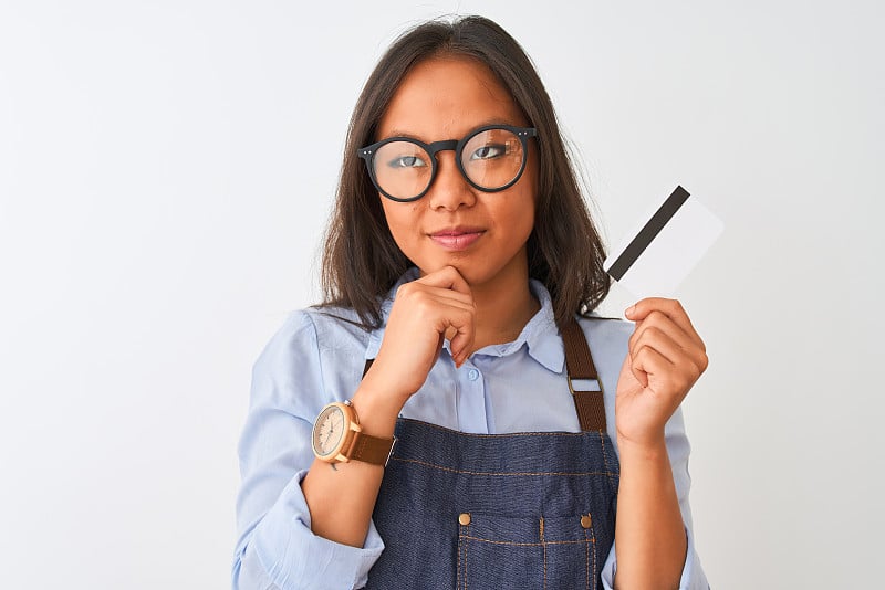 中国女店主戴着眼镜拿着信用卡越过孤立的白色背景严肃的脸思考问题，非常困惑的想法