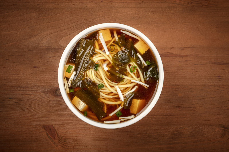 头顶上的照片是一碗味增汤，里面有豆腐、大葱、面条、裙带海藻和金木蘑菇，背景是深色的木头，有复制空间
