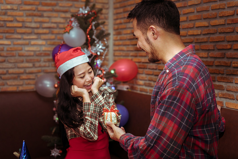 年轻男子惊喜小礼盒女友夫妇一起在圣诞派对与圣诞帽庆祝情人在家