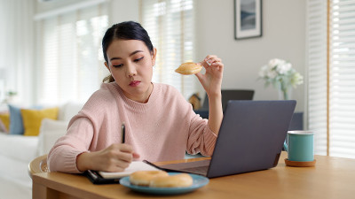 年轻迷人美丽的亚洲女性饿了吃甜甜圈，吃零食吃满嘴看电脑笔记本在家里在忙碌的工作，从家里多任务不健康的