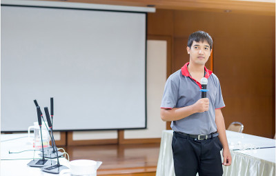 在会议室里，演讲者拿着麦克风，用白色的投影仪屏幕向听众展示他的食指
