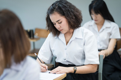 选择性焦点的青少年大学生坐在讲座椅上做期末考试和写在试卷上的答题卡在教室里。大学生在教室里穿制服。