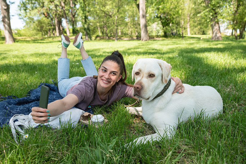 年轻美丽的女人躺在公园和她的宠物狗一只金色的拉布拉多寻回犬，用她的智能手机拍一张自拍，同时享受一个美