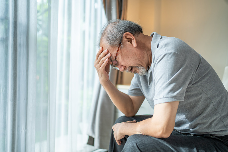一名成年的老年男子坐在家里的沙发上，头疼得厉害。压力大的老年男性因为可怕的偏头痛触摸他的太阳穴。医疗