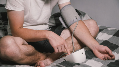 年轻白人男子在家测量血压，坐在床上在家测衣服。男性用医疗仪器血压计测量自己的血压。年轻时患高血压。