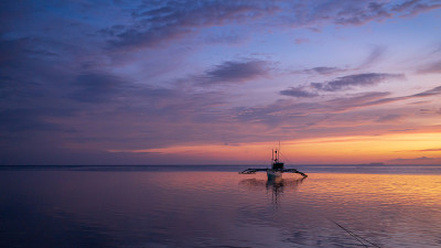 在紫色和橙色的天空下，夕阳照亮了Paliton海滩上的一艘小船