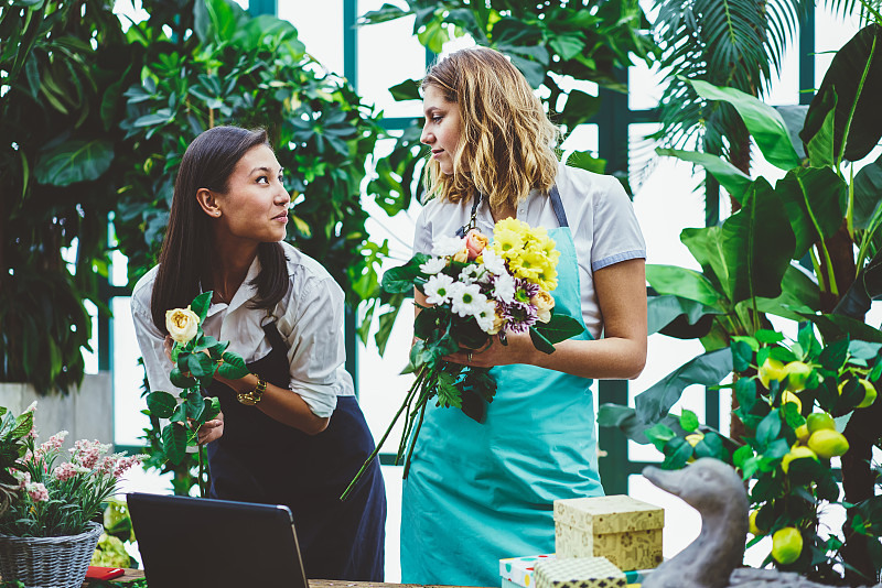 植物橙花店花店女同事合作创作花卉组合，系围裙的年轻女子协助花店专业店主制作花束供客户订购