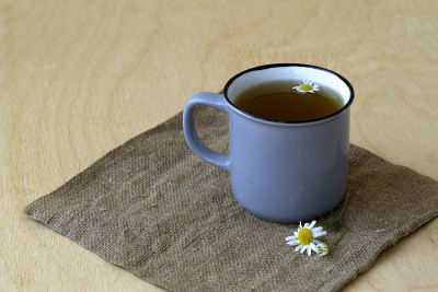 灰杯的洋甘菊茶，铺在麻袋布上。在一张木桌上放着一杯蓝色的热草药甘菊茶，在明亮的背景下放着鲜花和绿叶。