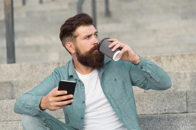 英俊的胡子男子放松手机和咖啡杯。在线生活。社交网络。现代生活在线交流。有智能手机城市空间背景的家伙。