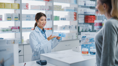 药店收银台:专业的亚洲女性药剂师推荐药物在一个美丽的白人女性客户的包装。