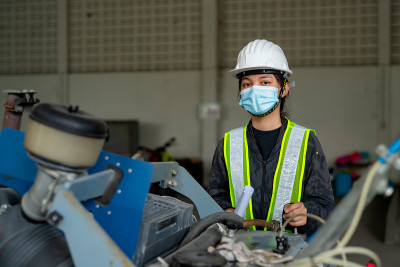 年轻的亚洲女工程师戴着医用口罩和白色安全帽在工地检查机器。