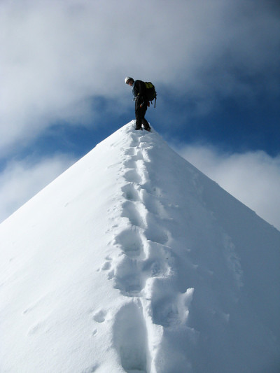 一个登山者，站在一个狭窄的、裸露的、有轨道向上的雪山山顶上