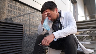 严肃的医生在医院上了很长时间班后正在楼梯上休息。疲惫的医生戴着口罩，白色制服和听诊器坐在诊所外面的楼