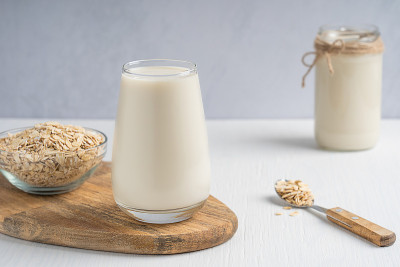 一杯奶油无乳制品素食燕麦牛奶与一碗薄片在木板上，勺子和罐子在白色的桌子上