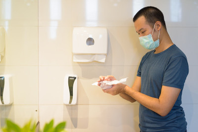 年轻亚洲男子戴口罩，并在浴室用纸巾擦干双手