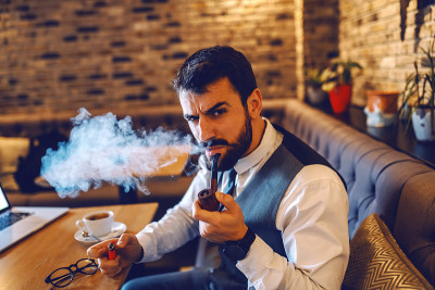 年轻认真有吸引力的白人商人穿着西装坐在咖啡馆，休息时抽着烟斗。
