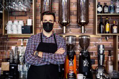 冠状病毒大流行期间的饮料服务。那个男人站在餐厅柜台前，穿着格子衬衫，系着围裙。一位女服务员双手交叉戴
