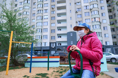 一个女孩坐在有公寓的高层建筑附近操场上的跷跷板上，她的脸上戴着医用口罩，防止病毒和灰尘