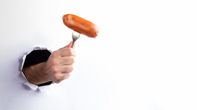男人的手拿着热煮香肠叉穿过破洞在白纸墙上。街头食品。垃圾食品。
