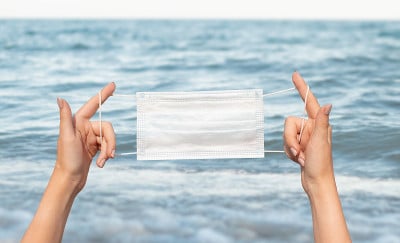 女性手中拿着医用防护口罩，背景是大海、大海。冠状动脉病毒感染流行期间的休息和旅行的概念。预防流感，烟