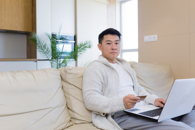 年轻快乐的亚洲男子使用笔记本电脑和信用卡在网上商店购物。男性坐在家里客厅的沙发上，在应用程序中输入服