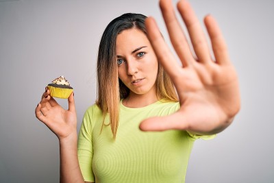 年轻美丽的女人与蓝色的眼睛吃甜蜜的巧克力纸杯蛋糕在白色的背景与开放的手做停止标志与严肃和自信的表情，