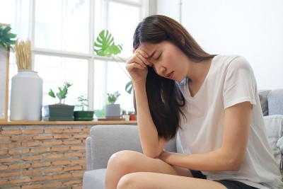 手搭在两鬓的年轻悲伤亚洲女孩坐在沙发上。由于生病和头痛，她感觉不太好。压力和疾病的概念。