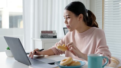 年轻迷人美丽的亚洲女性饿了吃甜甜圈，吃零食吃满嘴看电脑笔记本在家里在忙碌的工作，从家里多任务不健康的