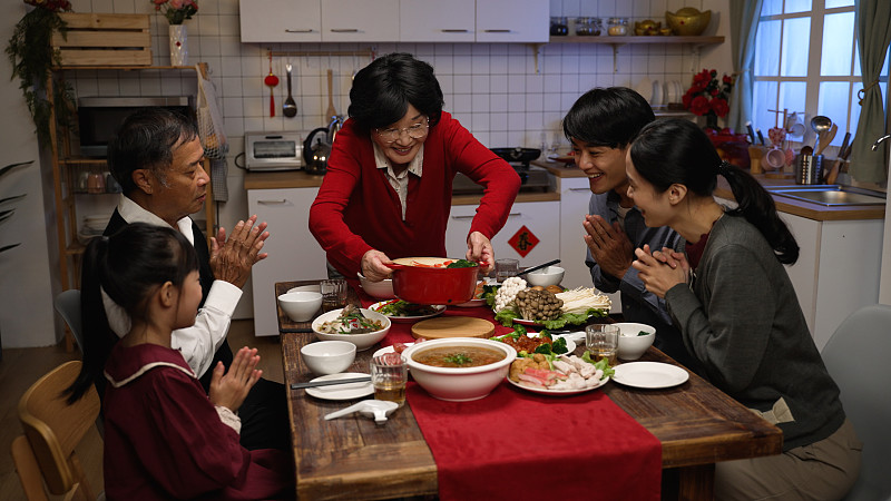 穿着红色衣服的快乐奶奶在餐桌上摆上美味的食物，她的家人为她鼓掌。为除夕的大餐做准备