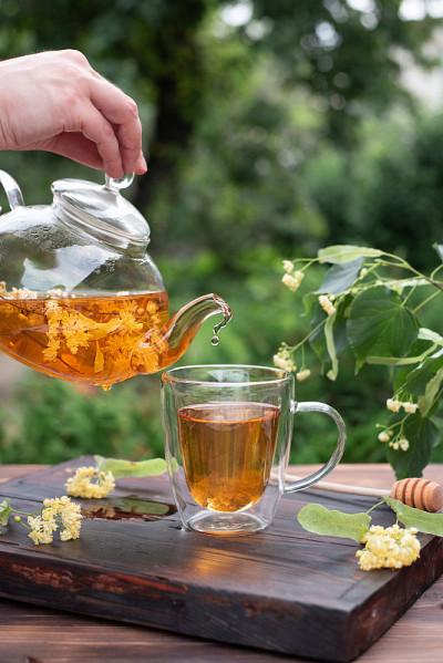 健康的花草茶，女性在花园里用手把菩提树茶从玻璃茶壶倒进杯子里。