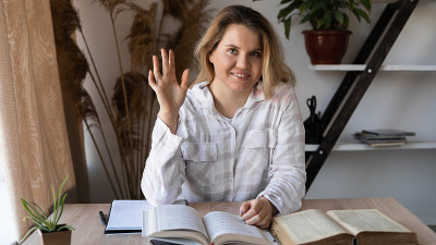 一位年轻的女教授坐在靠窗的木桌旁，桌上放着书本和课本。一位女士正准备召开视频会议，她对着镜头挥挥手。