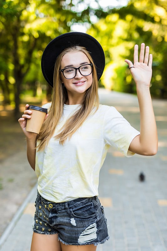 一名年轻女子端着一杯咖啡在户外向别人打招呼