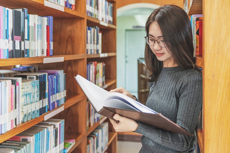 回到学校教育知识学院的大学理念，美丽的女大学生抱着她的书微笑着站在图书馆，学习和教育的理念