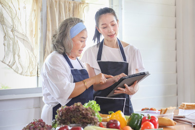 一名退休年龄的亚洲老年妇女花时间学习如何用数码平板电脑和互联网烹饪健康食物，她的女儿用各种蔬菜