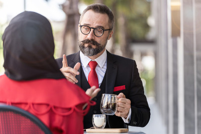 一位穿着西装的高级男士坐在户外的咖啡桌上与人交谈。