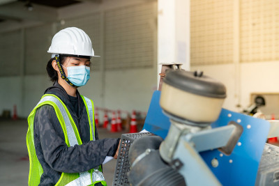 年轻的亚洲女工程师戴着医用口罩遮住嘴，戴着白色安全帽检查施工区内的大型地板切割机。