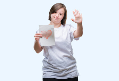 年轻的成年妇女与唐氏综合症拿着红心卡片在孤立的背景与开放的手做停止标志与严肃和自信的表情，防御手势