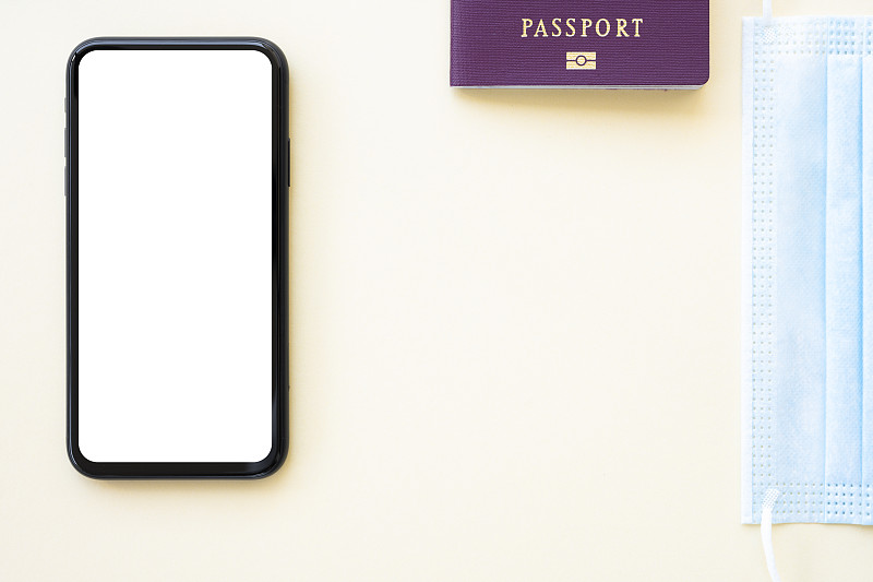 在奶油色的背景上，放着一本护照、一个口罩和一部白色屏幕的智能手机，平铺着复印空间。在新冠肺炎大流行期